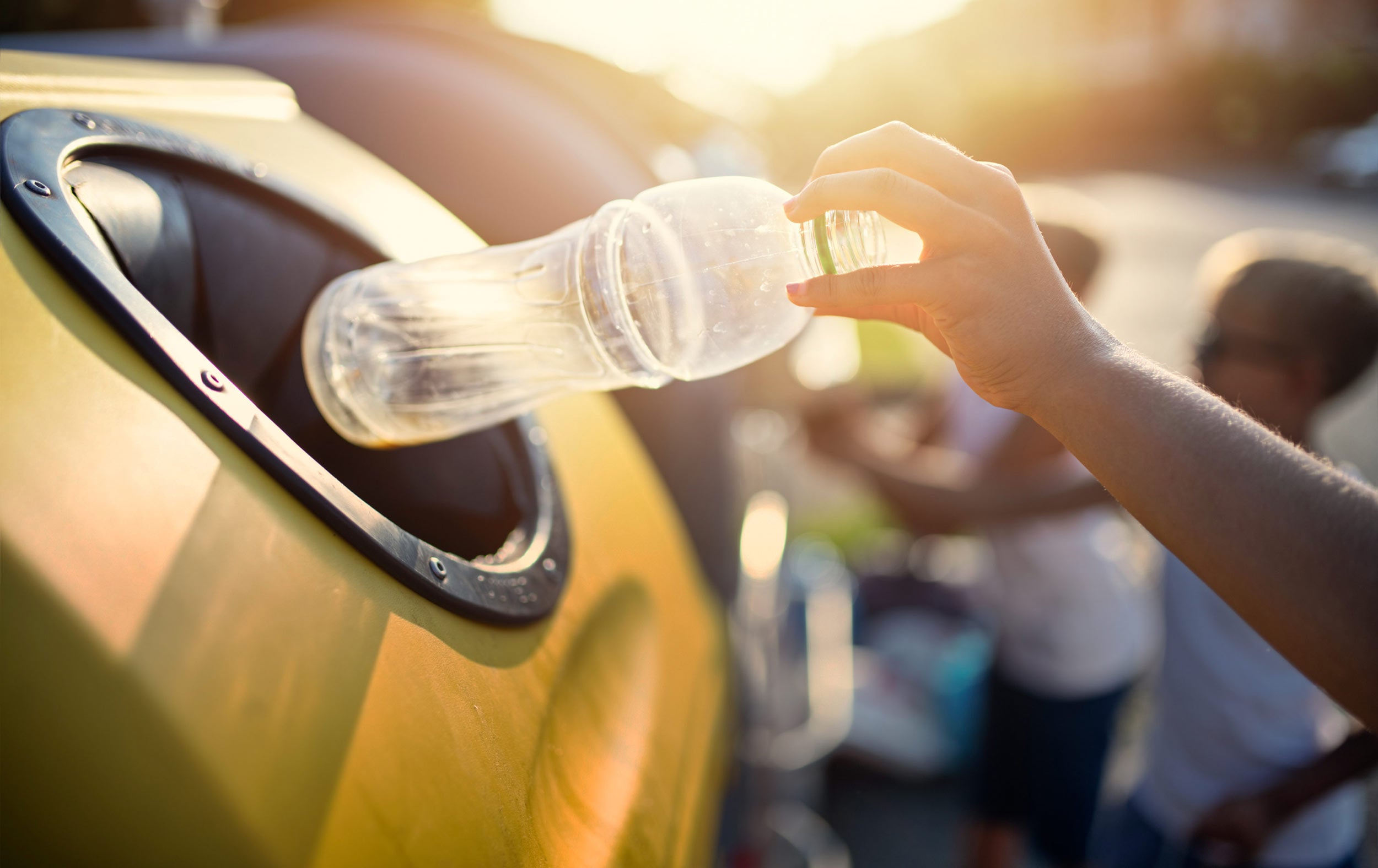 hand placing plastic bottle into a large yellow outdoor bottle recycling bin